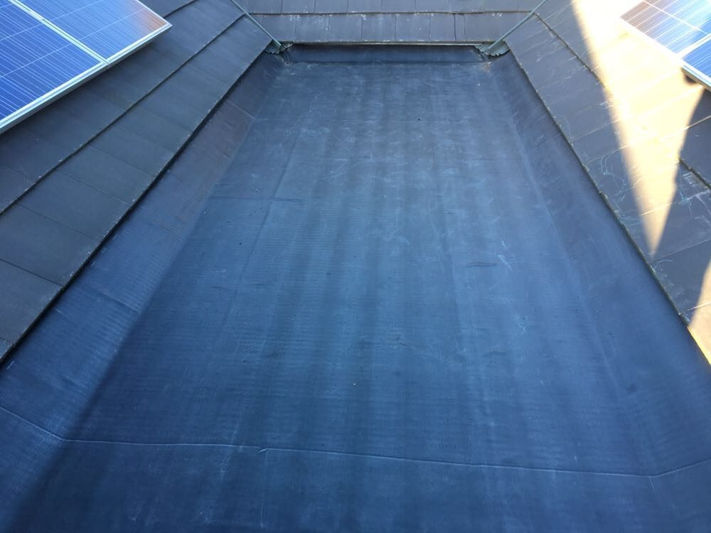 Close view of a dark waterproofed roof valley between slate-covered roof slopes, with solar panels visible at the upper edges