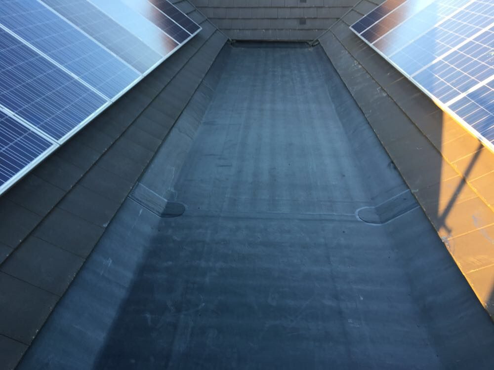 Completed dark waterproofing membrane forming a central roof valley, with slate courses and solar panels visible on both sides