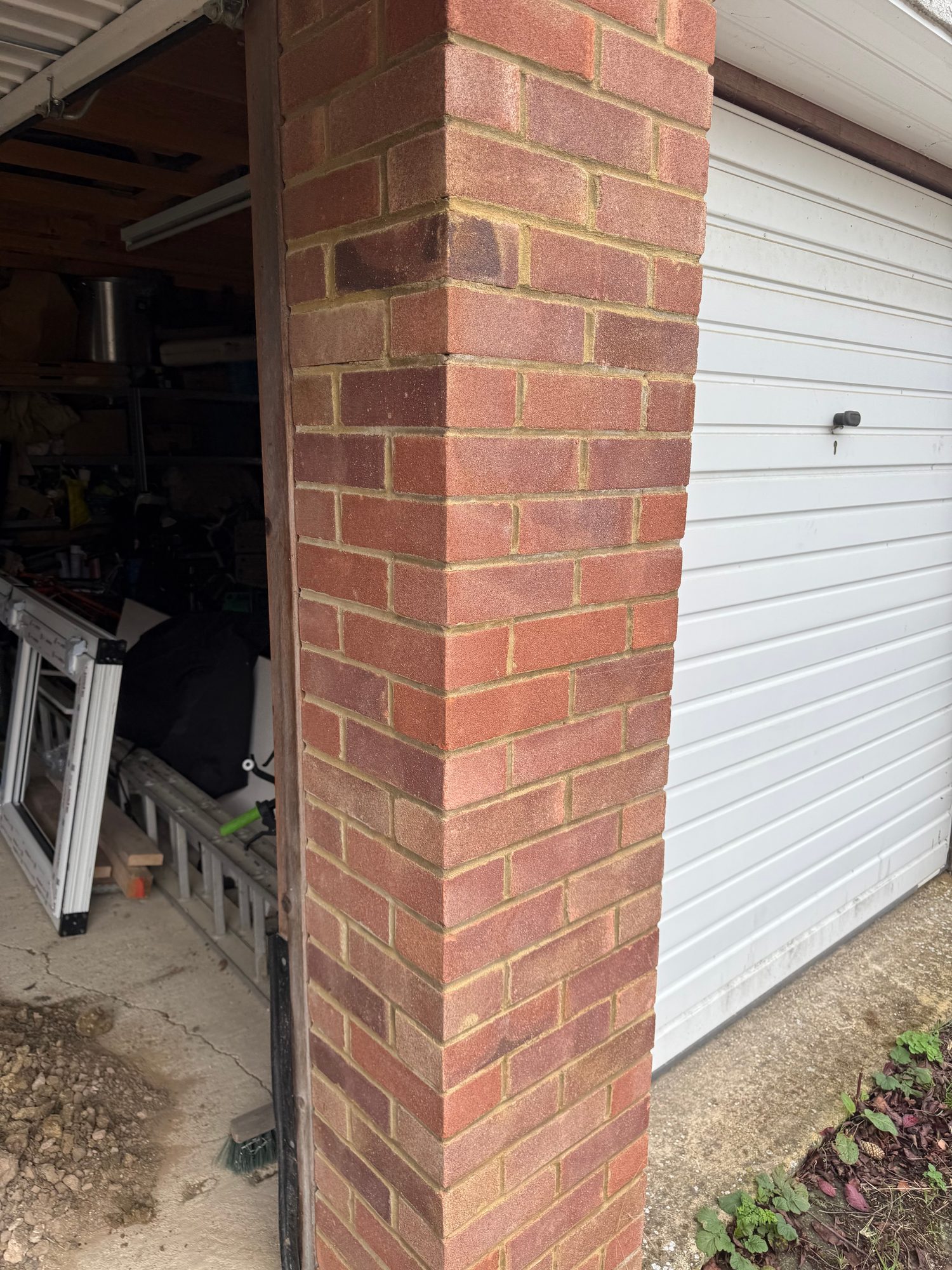 Exterior view of the garage showing a red brick corner, white roller garage door and the open garage entrance with stored tools and materials visible inside