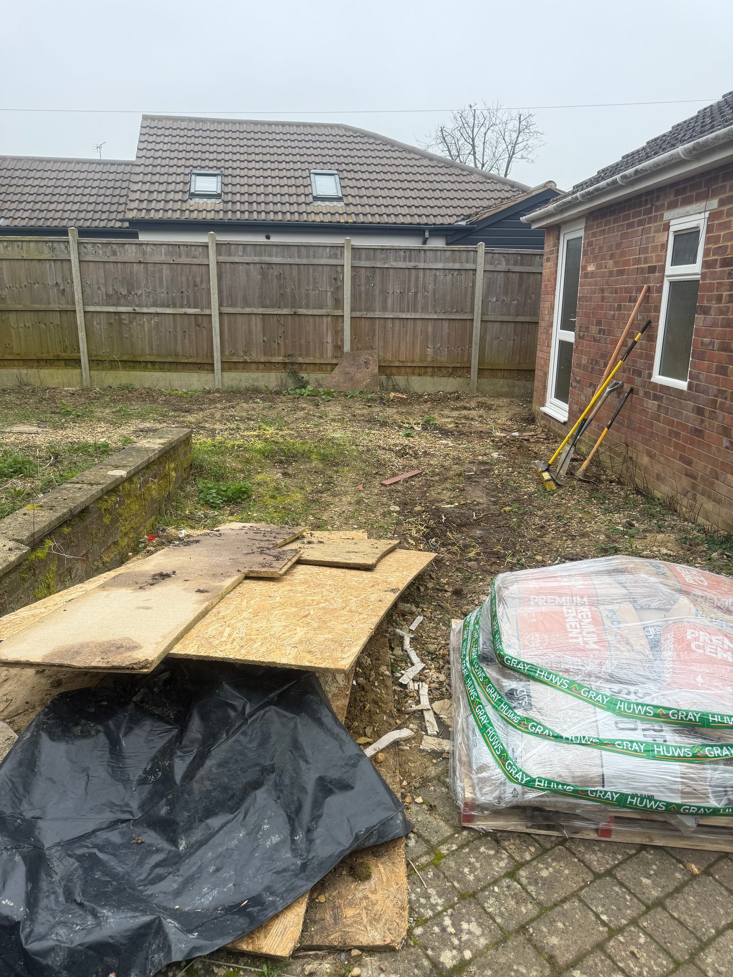 Rear garden area before renovation, showing bare soil, paving, timber boards, building materials and garden tools beside the brick extension