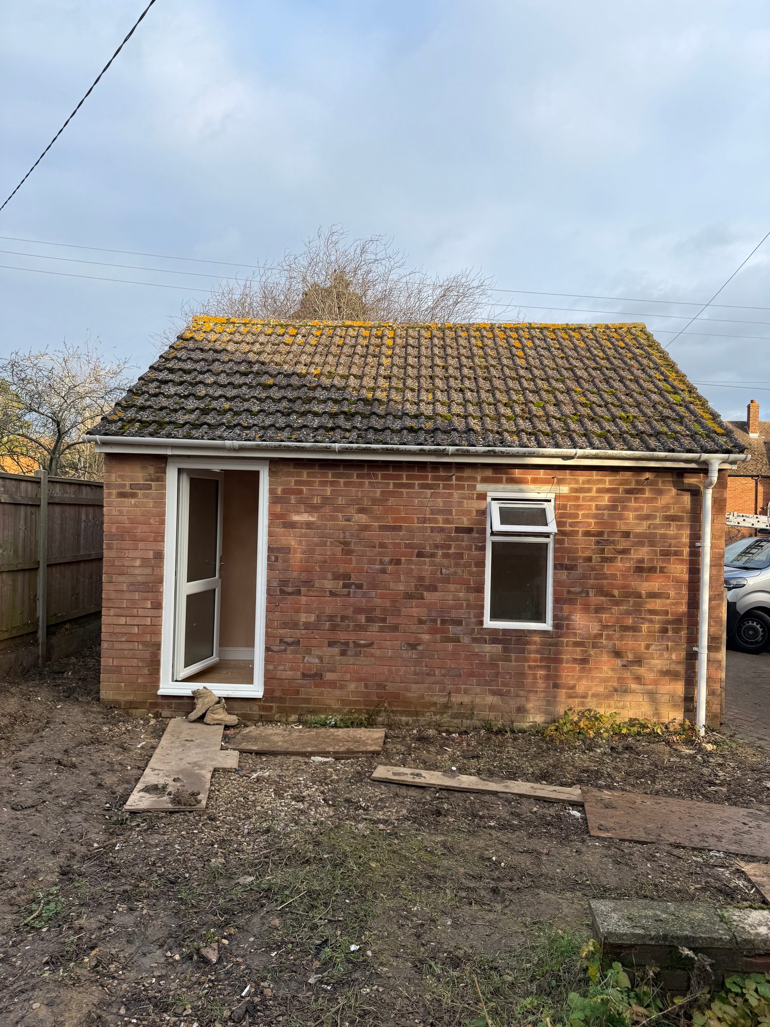 ear elevation of the brick building before renovation, showing the tiled roof, white-framed door and window, guttering and an unfinished ground area with timber boards