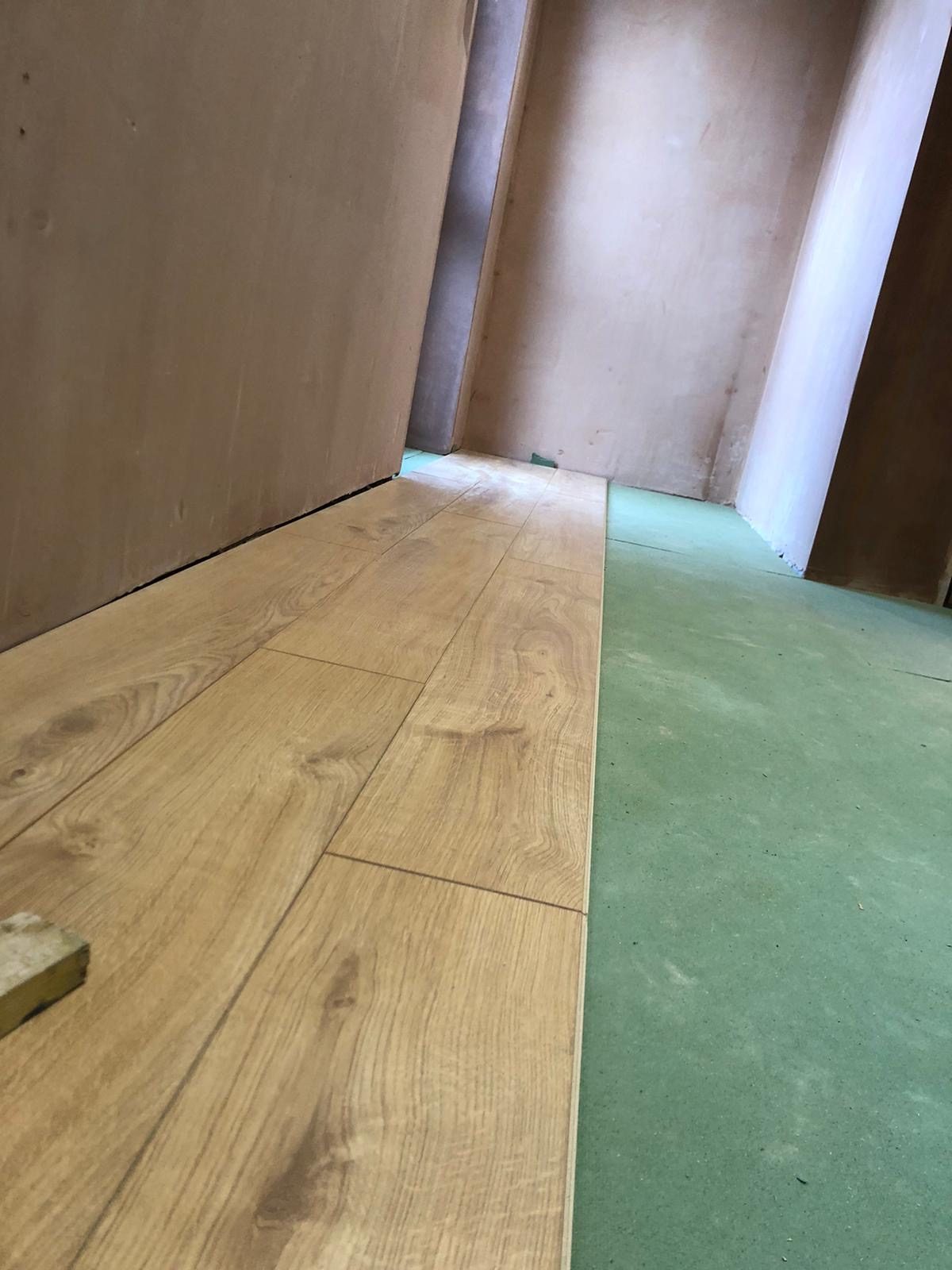 Partially installed wood-effect flooring runs along the hallway, with the remaining green floor surface visible beside it and unfinished door areas in the background