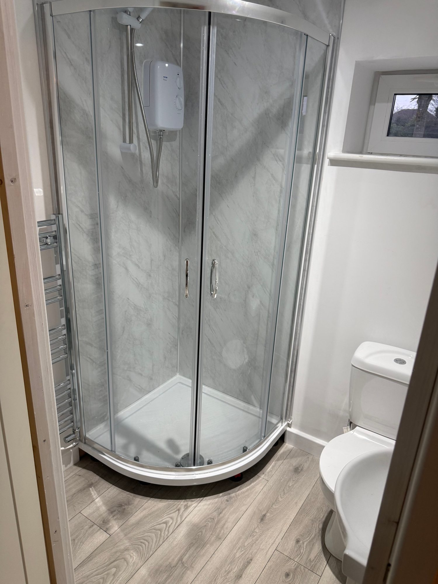 Finished bathroom with a curved glass shower enclosure, electric shower, white toilet, chrome towel radiator and light wood-effect flooring