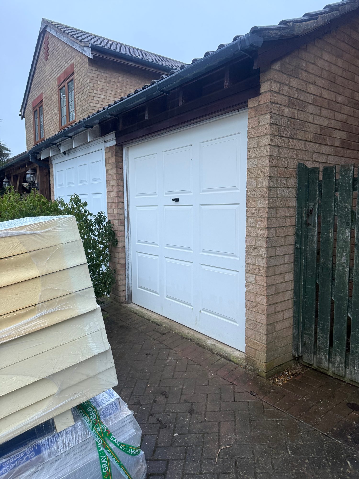 Two white garage doors beneath a tiled roof, with dark roofline timber detailing and wrapped building materials visible in the foreground