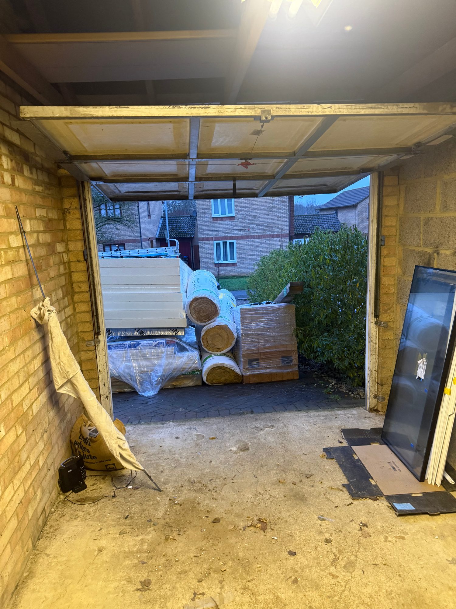 Interior view of a garage with the segmented door open, showing a concrete floor, exposed brick and blockwork walls, building materials stored outside and materials leaning against the right-hand wall