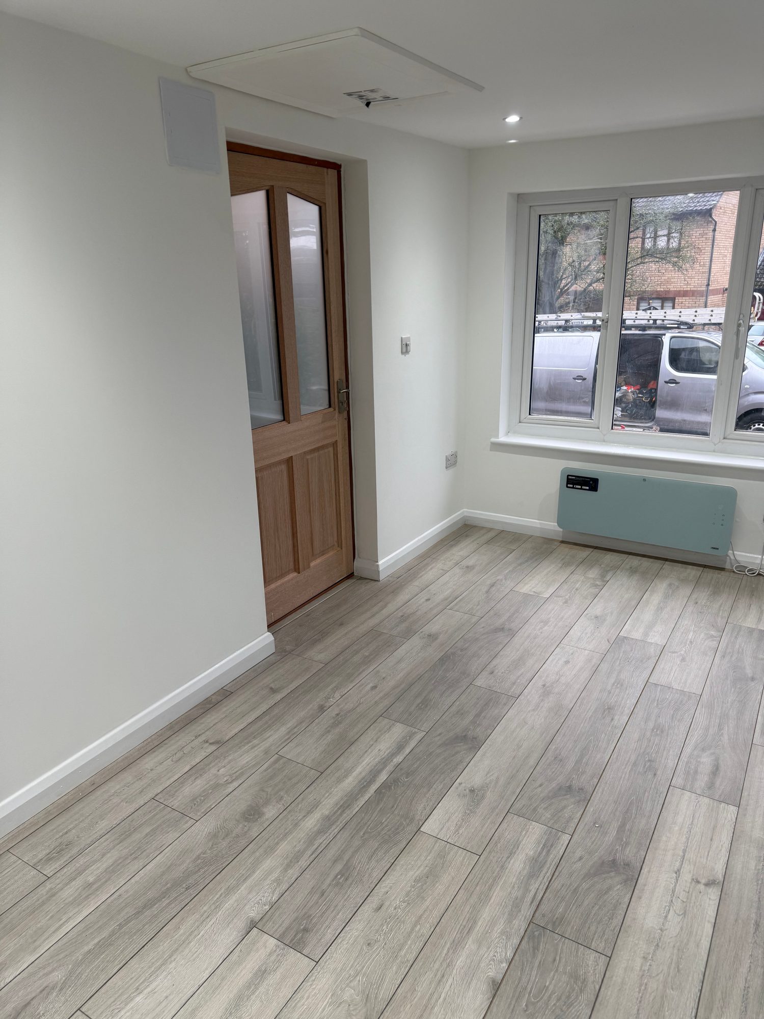 Finished interior room with light painted walls and ceiling, grey wood-effect flooring, recessed ceiling lights, a wall-mounted heater, white-framed window and timber internal door
