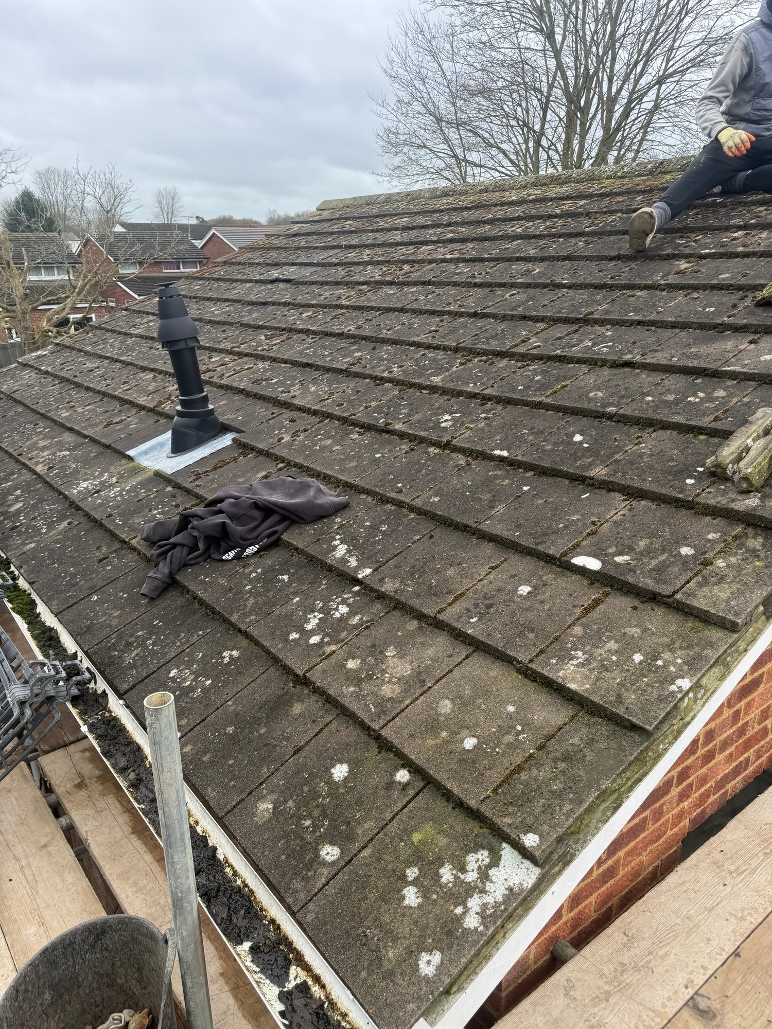 Pitched roof covered with concrete tiles showing widespread moss, lichen and weathering, with a roof vent and a worker partially visible at the upper right