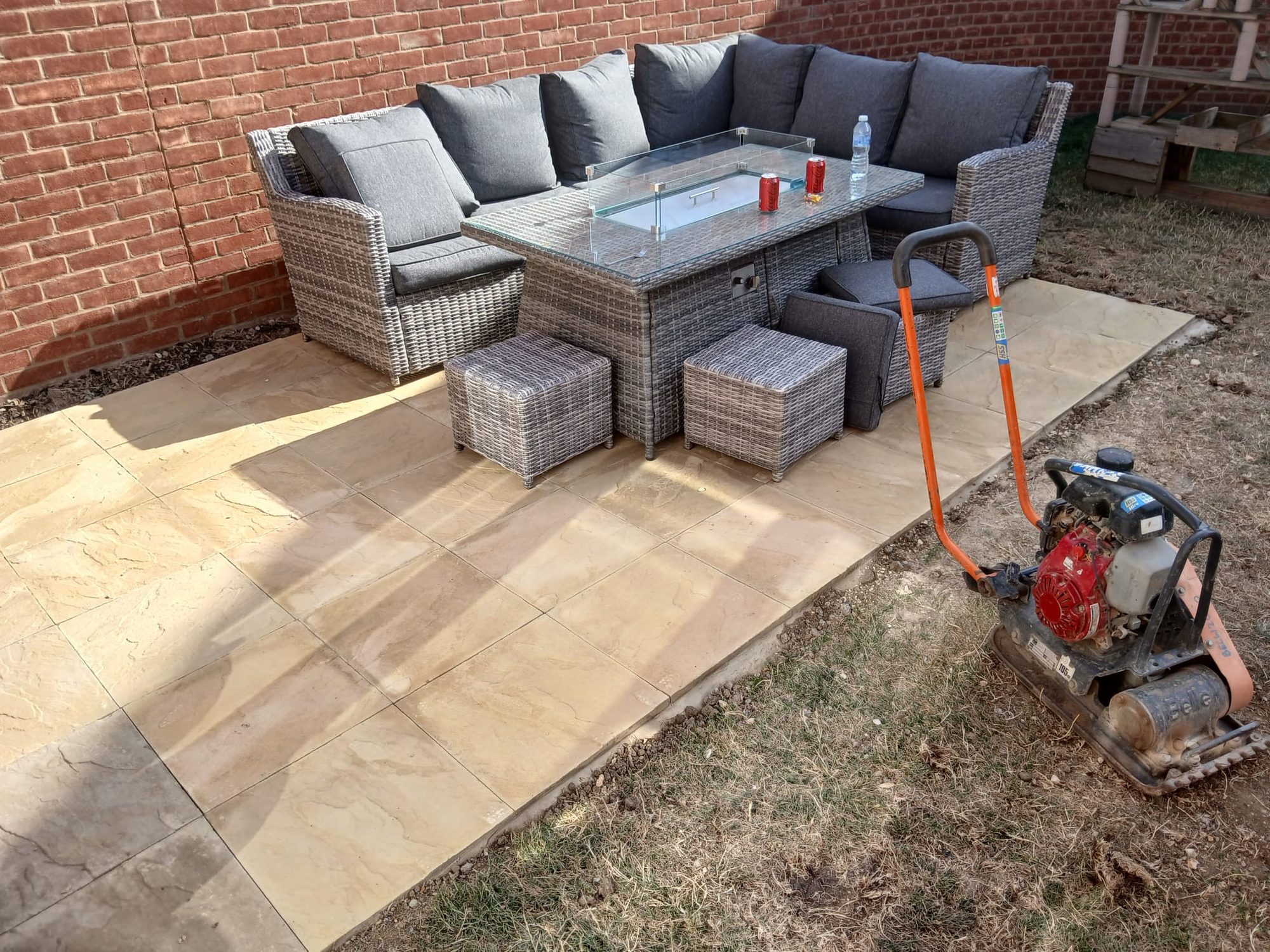 Buff-coloured patio with outdoor seating beside a brick wall and a plate compactor on the surrounding ground