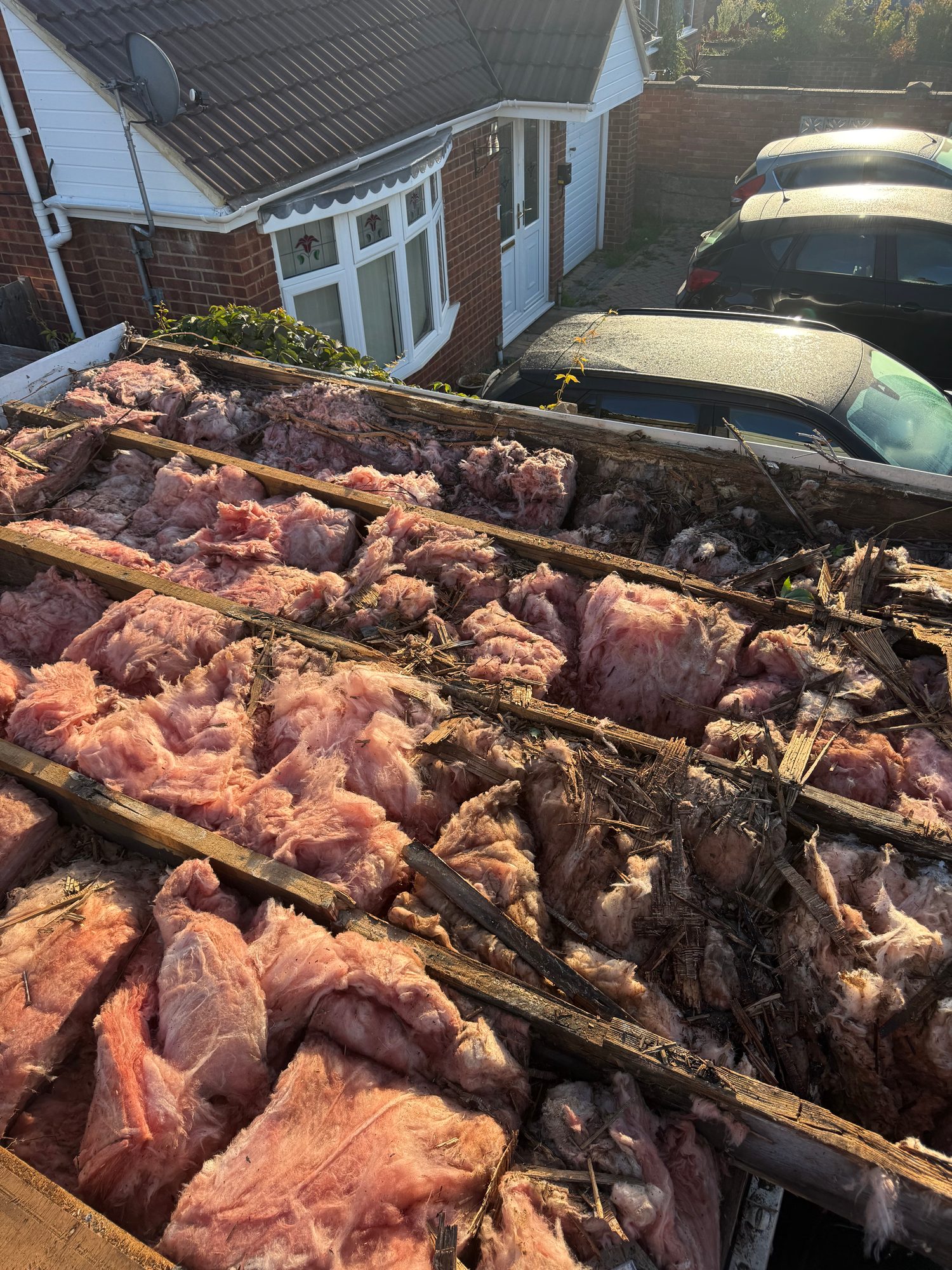 Partially stripped roof showing exposed pink insulation, timber battens and roof materials, with a house, parked cars and garden wall visible in the background