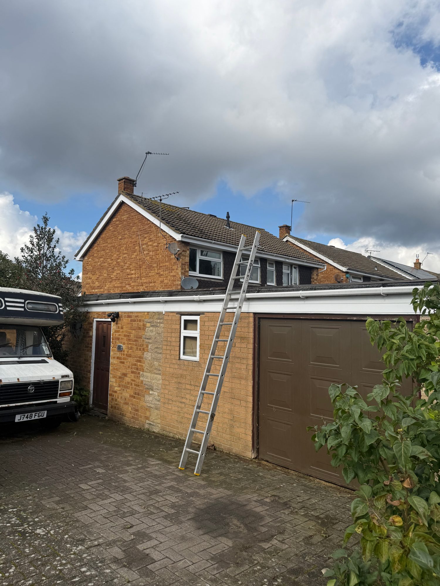 Exterior view of a brick property before works, showing the garage, tiled roof, driveway and an extension roof with a ladder in place