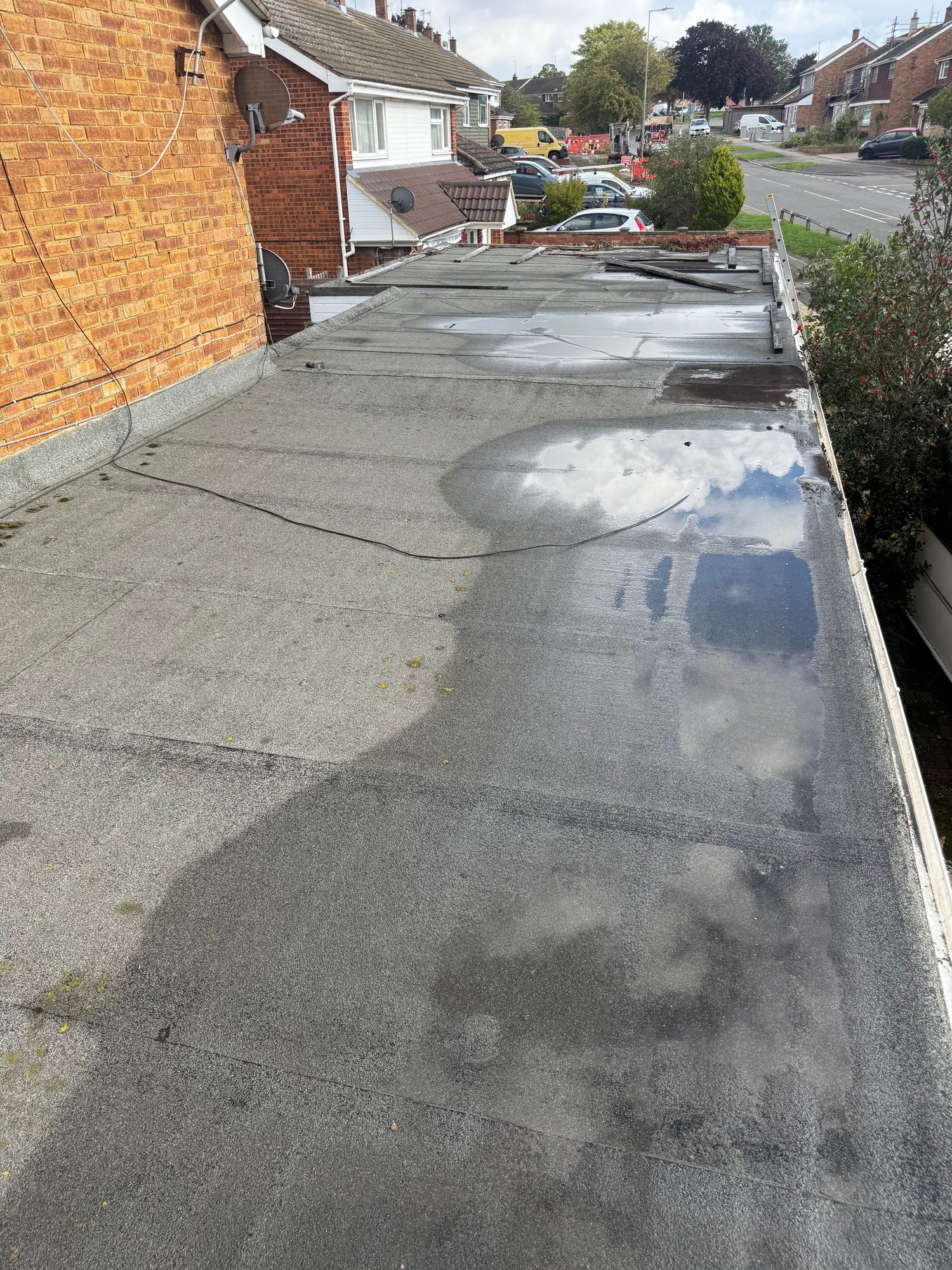 Flat roof viewed from above with visible seams, patches of standing water and sections of loose or damaged roof covering near the far edge, with neighbouring houses and a road in the background